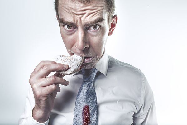 A man who has jam on his tie and sugar on his face is biting a donut and looking at us with a worried expression.