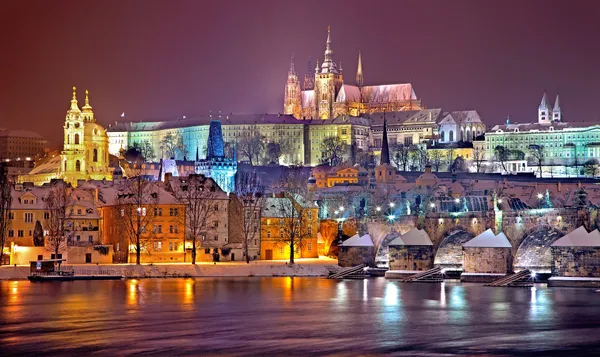 Old buildings in Prague that are lit up at night and seen from across a river.