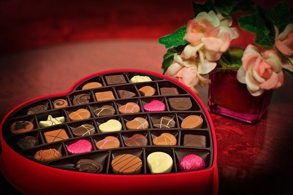 An open heart-shaped box of chocolates next to flowers.