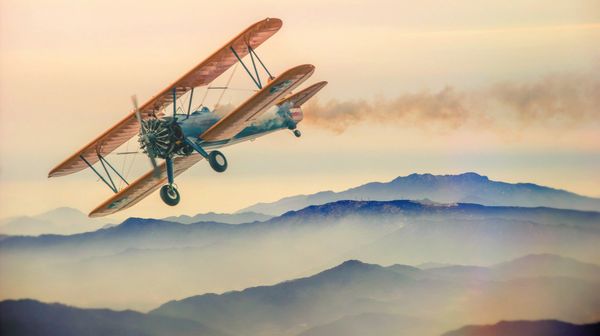 A biplane in the sky over mountains and clouds. There is smoke coming from the biplane.