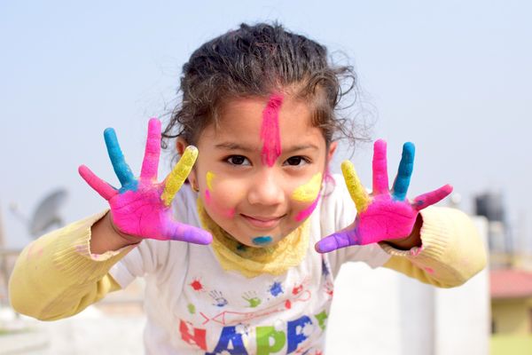 A girl with bright paint on her hands and face. She is spreading the fingers of her hands widely.