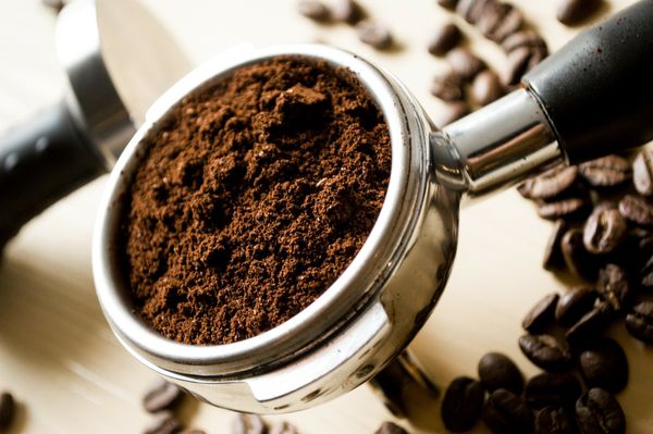 Ground coffee in an espresso machine portafilter. There are coffee beans in the background.