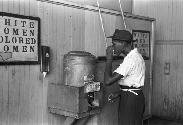a black man drinking from a water cooler next to a sign that says 'colored'