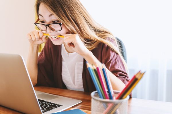 A woman who is wearing glasses and biting a pencil is staring at a computer, as if she is studying hard.