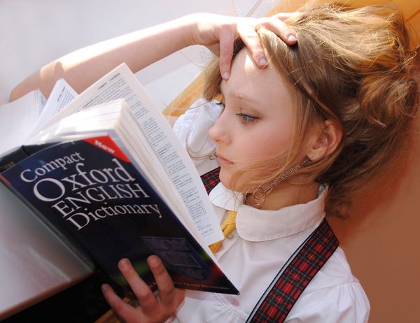 A girl is lying down and reading a dictionary. Her hand is on her head.