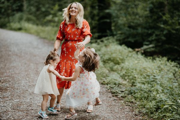 A woman with blond hair is wearing a red dress and holding the hands of two little girls. They are on a path in a forest.