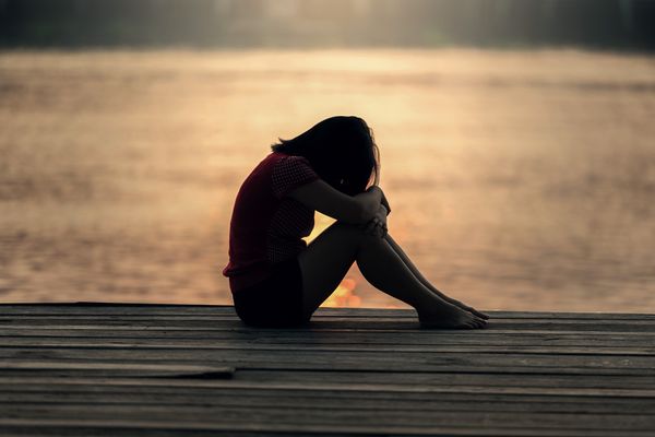 A girl is sitting on a pier next to water with her head down resting on her arms.