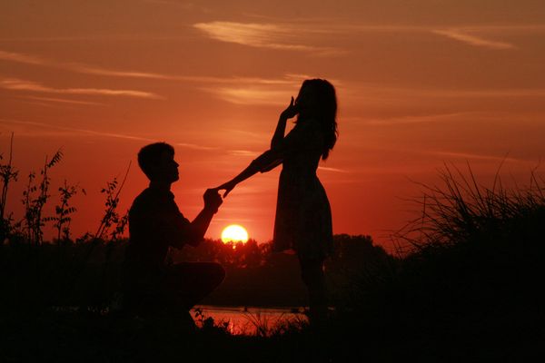 A man, kneeling, proposing to a woman, with the sun setting in the background.