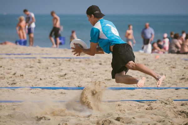 A man in sportswear diving to catch a frisbee on a beach. In the background we can see people relaxing by the ocean.