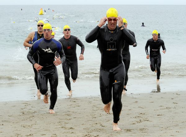 Men in wetsuits, bathing caps, and swimming goggles are emerging from the ocean onto the beach. They look like they are competing in a race.