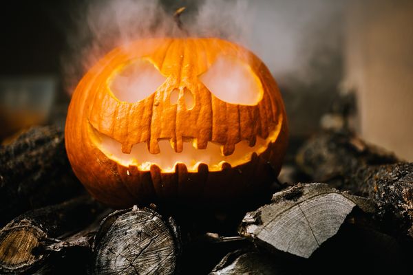 A jack o'lantern with smoke coming out of its eyes and mouth is on some black logs.