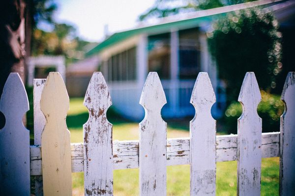 A worn white-picket fence with a house in the background.