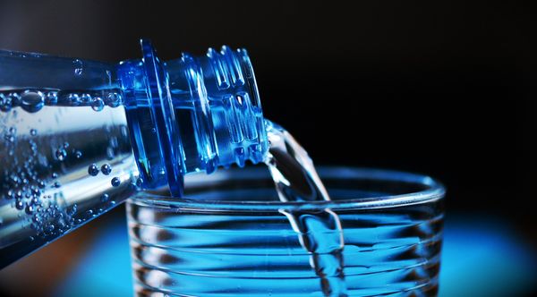 Water is pouring from a plastic bottle into a drinking glass that it is resting on. There is a blue tint to the image.