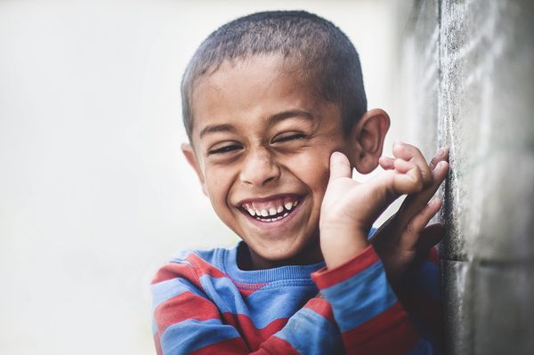 A smiling boy with his hands near his face and a wall on his left.