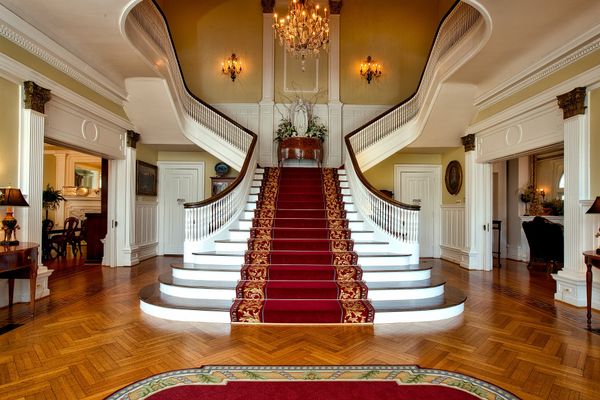 Inside the entrance of a mansion, there is a staircase with red carpeting and a hardwood floor.