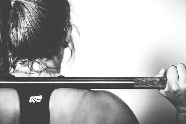 A view from behind of a weightlifting bar resting on a woman's neck and shoulders. Her hair is tied up.
