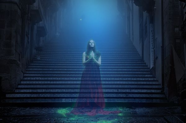 A ghostly woman wearing a dress and standing in front of a long staircase. She appears to be praying, and she is partially see-through.