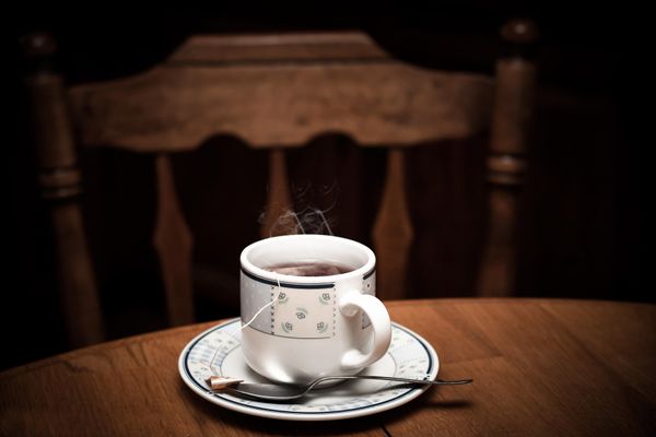 A steaming cup of tea on a saucer with a teaspoon. It's sitting on a wooden table next to a wooden chair in a dark room.